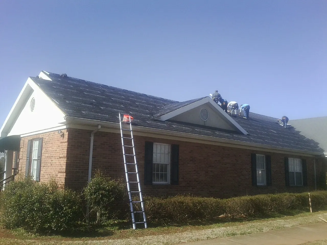 Crew tearing shingles off a brick ranch roof with a ladder set — Metal Roof Repair work by our crew in Crosby