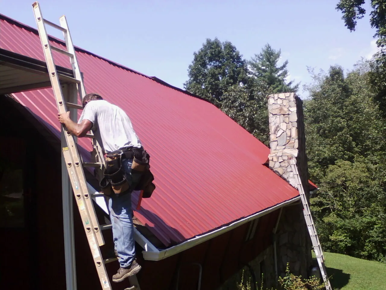 Roofer on a ladder beside a red metal roof and stone chimney — Soffit & Fascia Repair by Roofing Brothers in Crosby